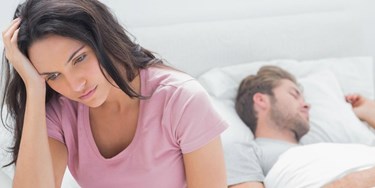 Woman on bed looking sad as man lays asleep beside her